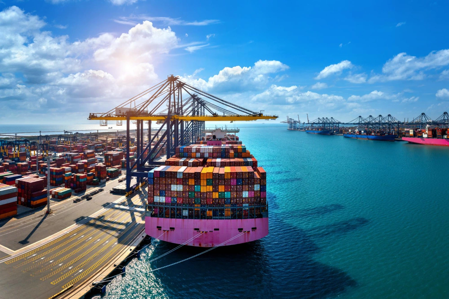 Large cargo ship loaded with colorful containers docked at a bustling port terminal under clear skies, with cranes and container stacks visible in the background.